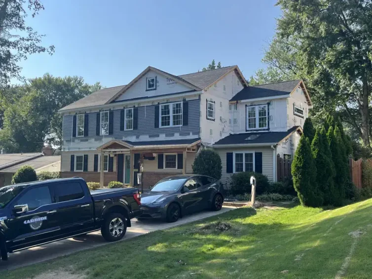 Two-story house under renovation with Tyvek wrap on the upper level, blue shutters, and two cars in the driveway.